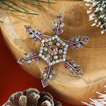 Load image into Gallery viewer, Decorative snowflake brooch with pink and clear gemstones on a wooden surface with pine cones and branches.